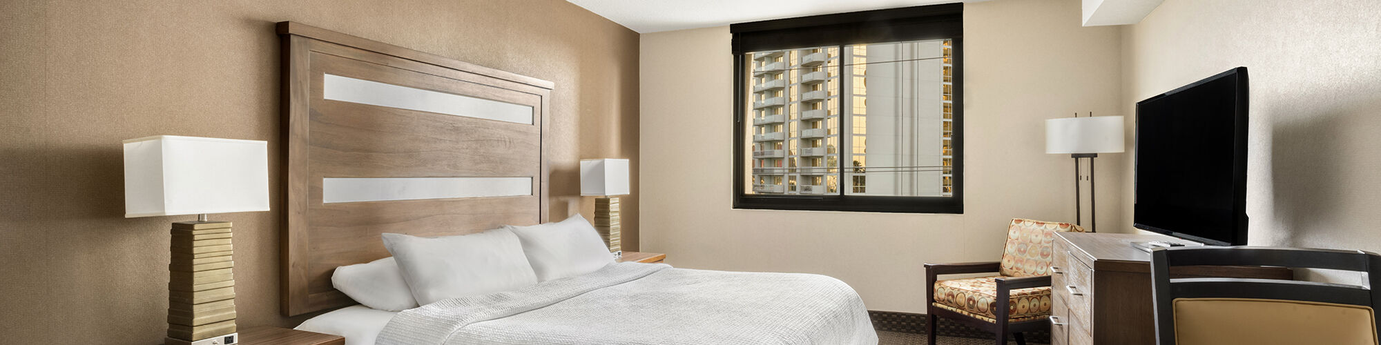 The image shows a hotel room with a neatly made bed, two bedside tables with lamps, a TV, a chair, a dresser, and a window with a city view.