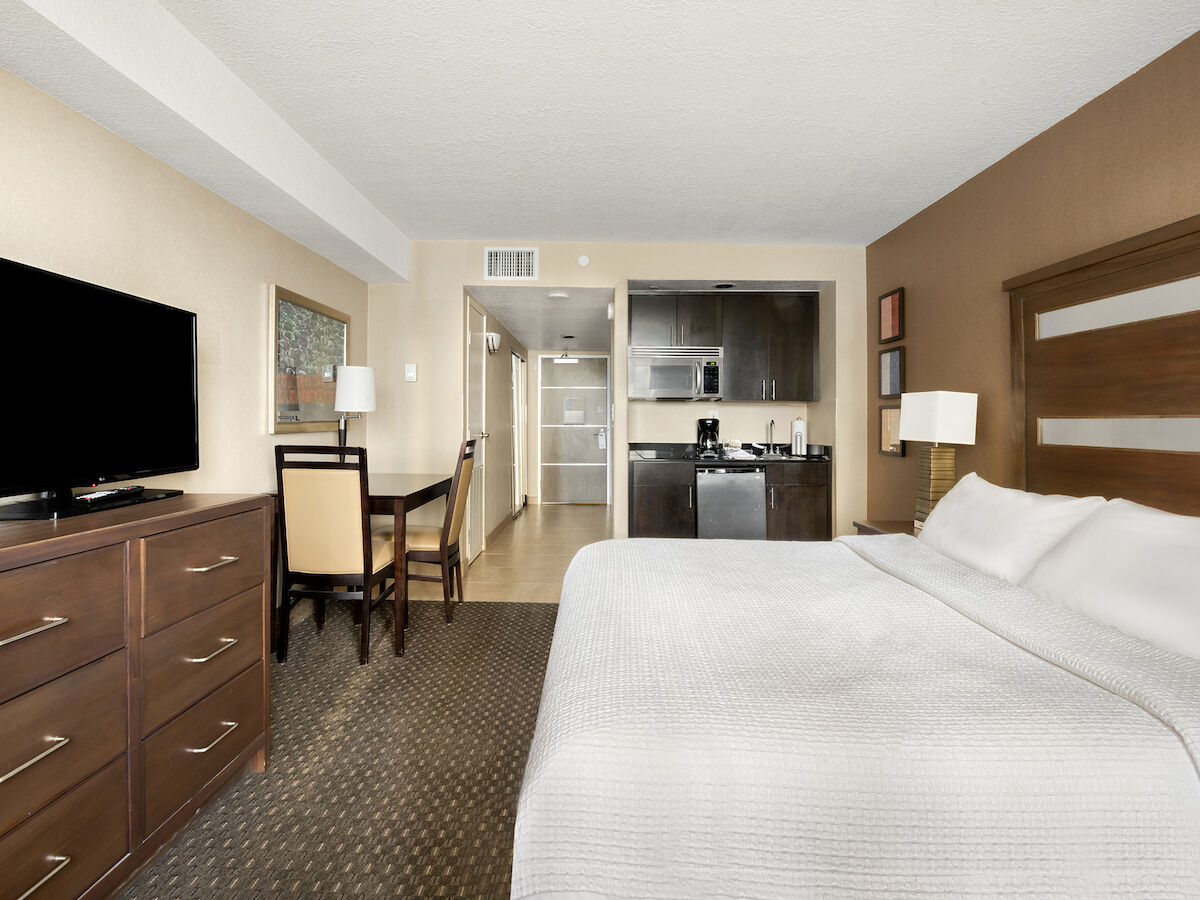 A clean, modern hotel room with a king-size bed, TV, dresser, desk, chairs, and kitchenette in the background.
