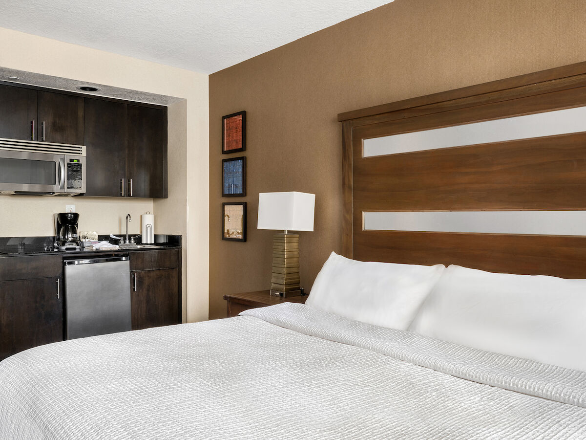 A modern hotel room features a bed, nightstand with a lamp, and a kitchenette equipped with a microwave, fridge, coffee maker, and utensils.