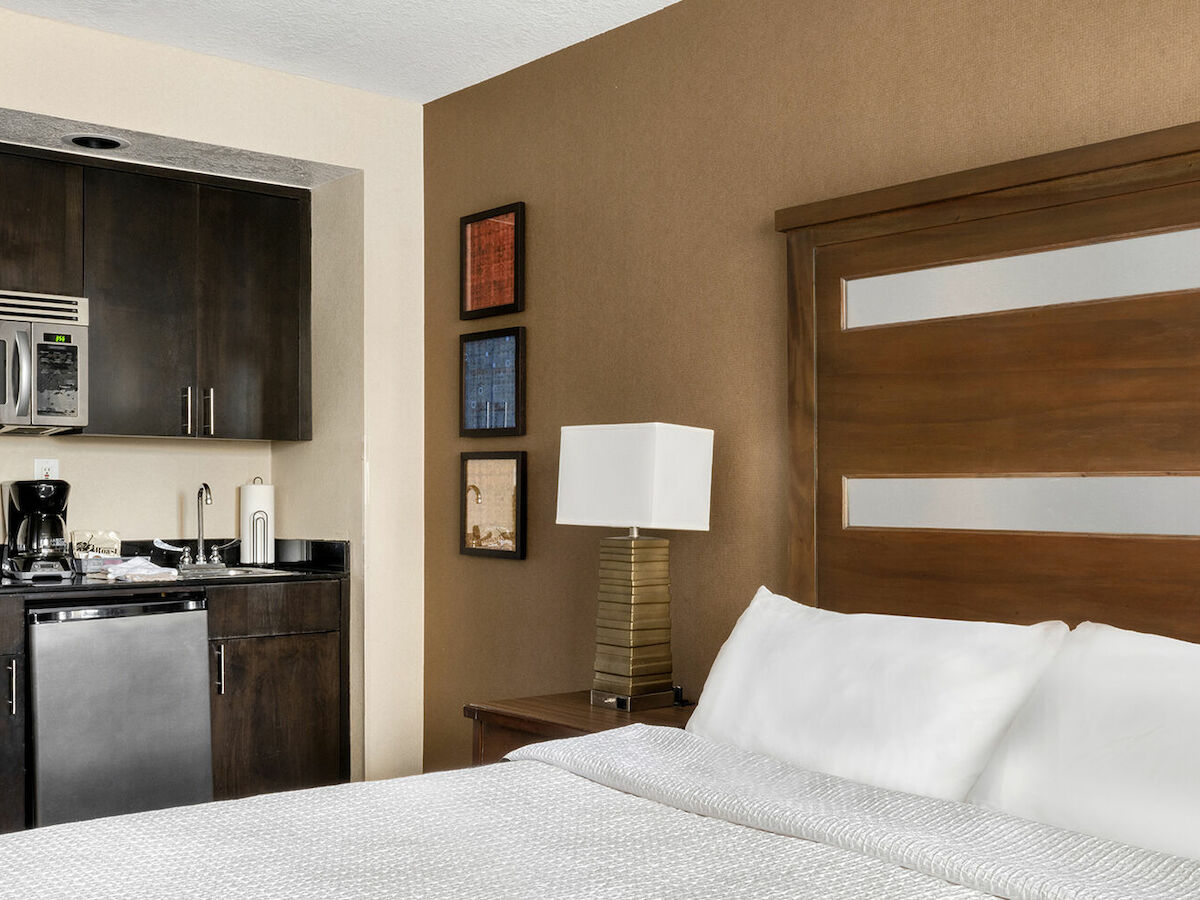 A hotel room with a bed, nightstand, lamp, and a small kitchenette with a microwave, coffee maker, sink, and cabinets. Artwork decorates the wall.