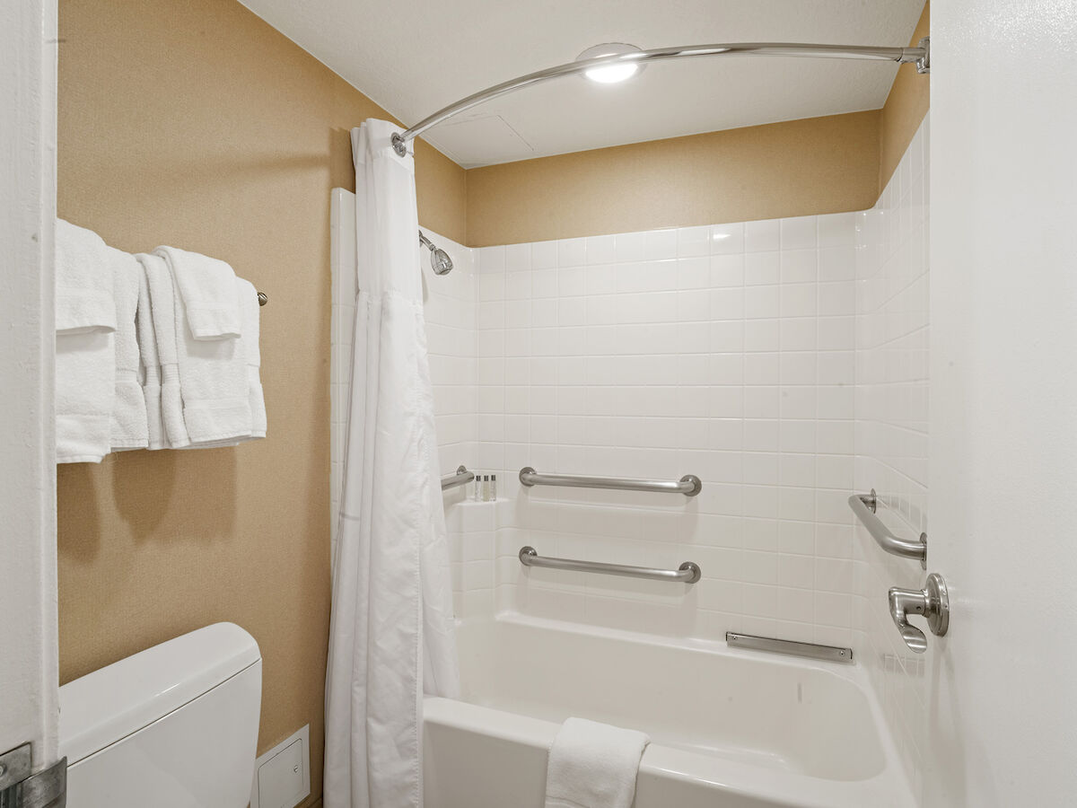A bathroom with a white bathtub, shower curtain, grab bars, and wall-mounted towels. The walls are beige, and there's an overhead light.