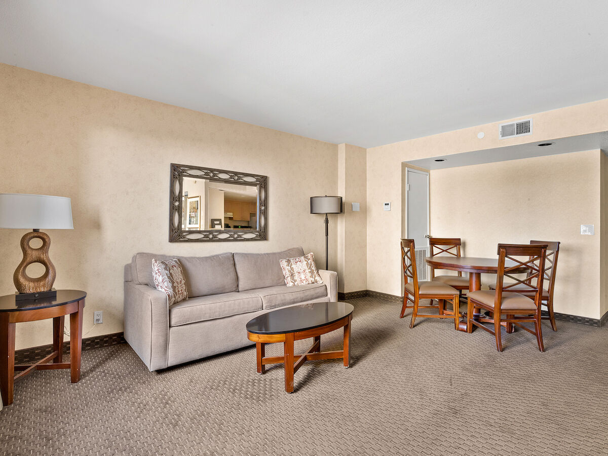 A cozy hotel room with beige walls, a couch with pillows, a coffee table, a mirror, a lamp, and a dining table with four chairs.
