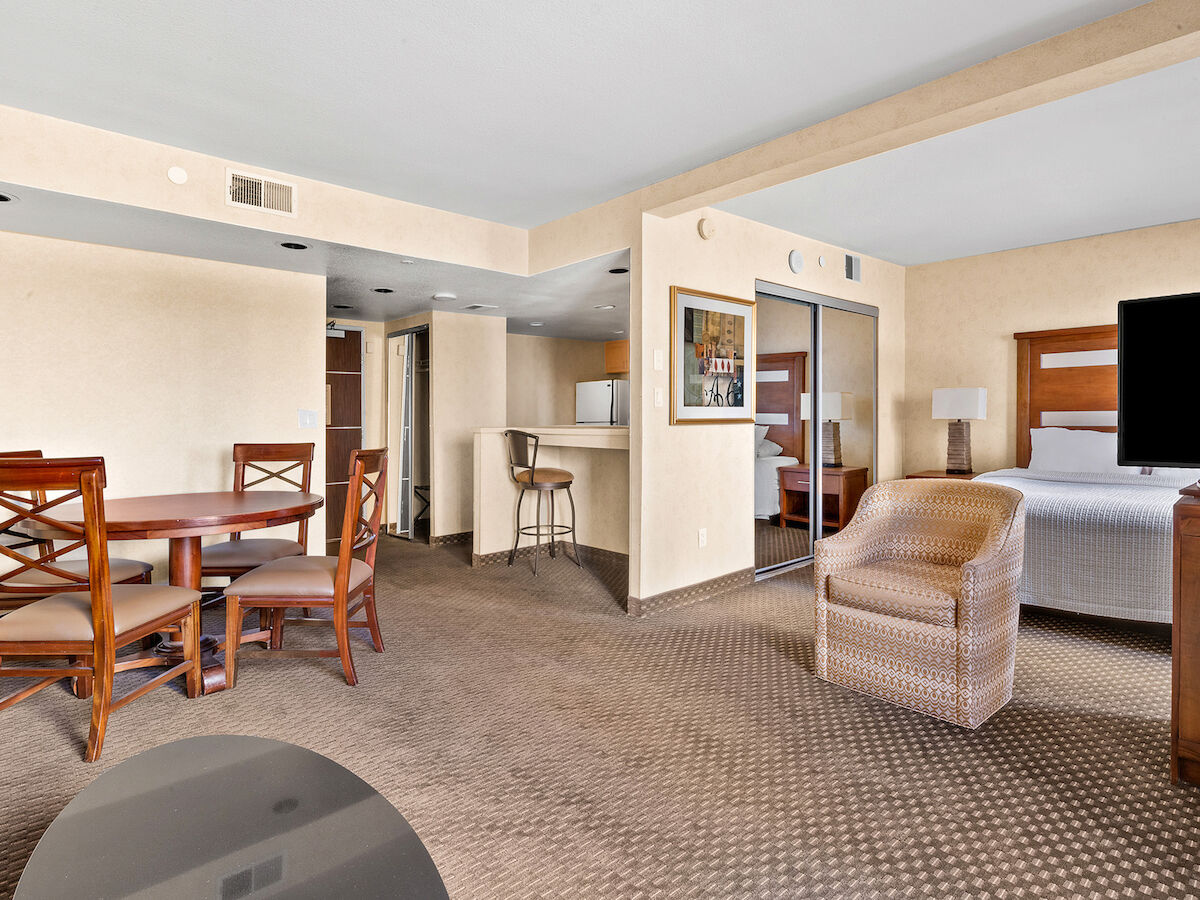 This image shows a hotel suite with a round dining table and chairs, a cushioned chair, a TV, and a bed with side tables and lamps.