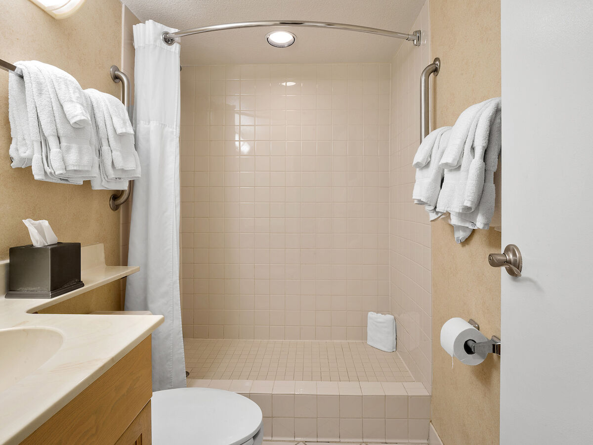 A clean bathroom with multiple white towels, a shower with a curtain, a sink with tissues, and a toilet. The floor and walls are tiled.