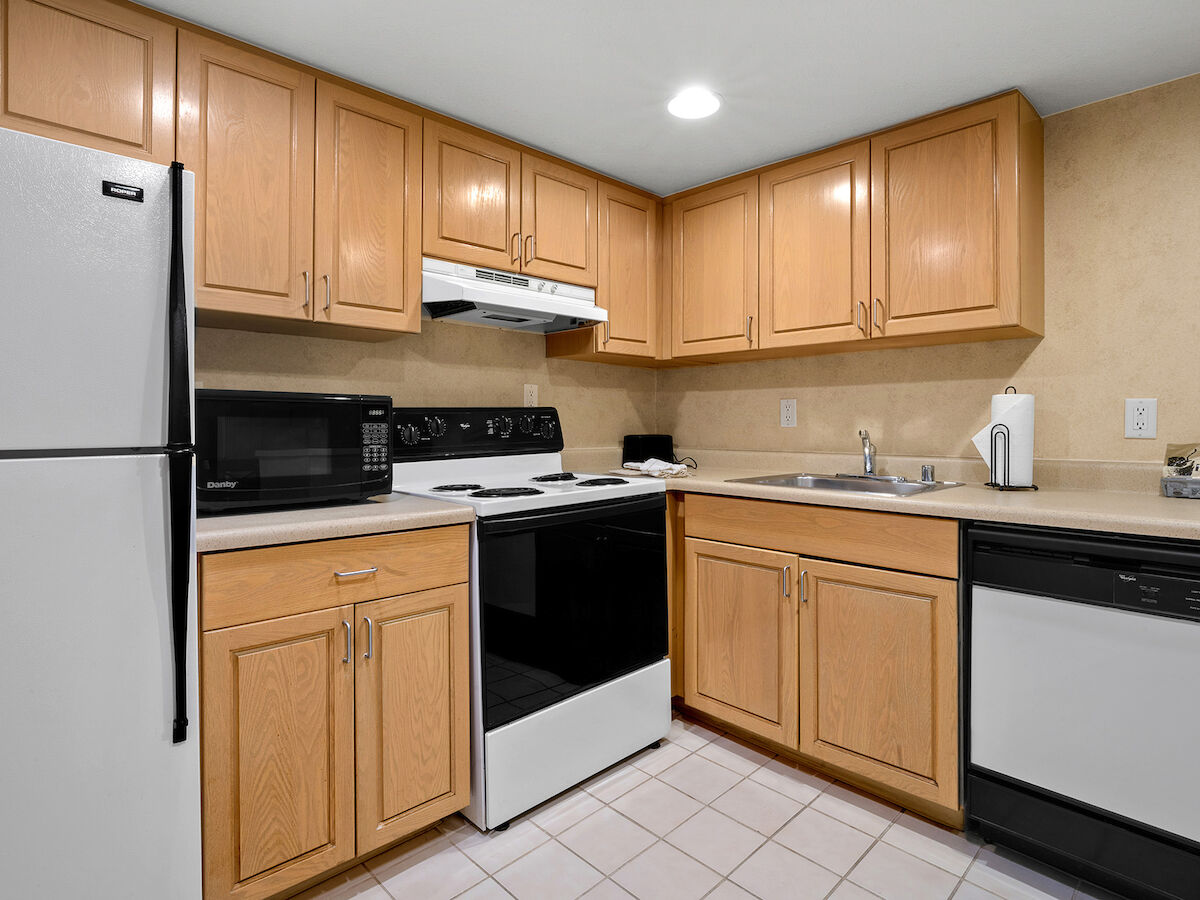 A kitchen with wooden cabinets, white appliances, and tiled flooring; includes a fridge, stove, microwave, sink, and dishwasher.