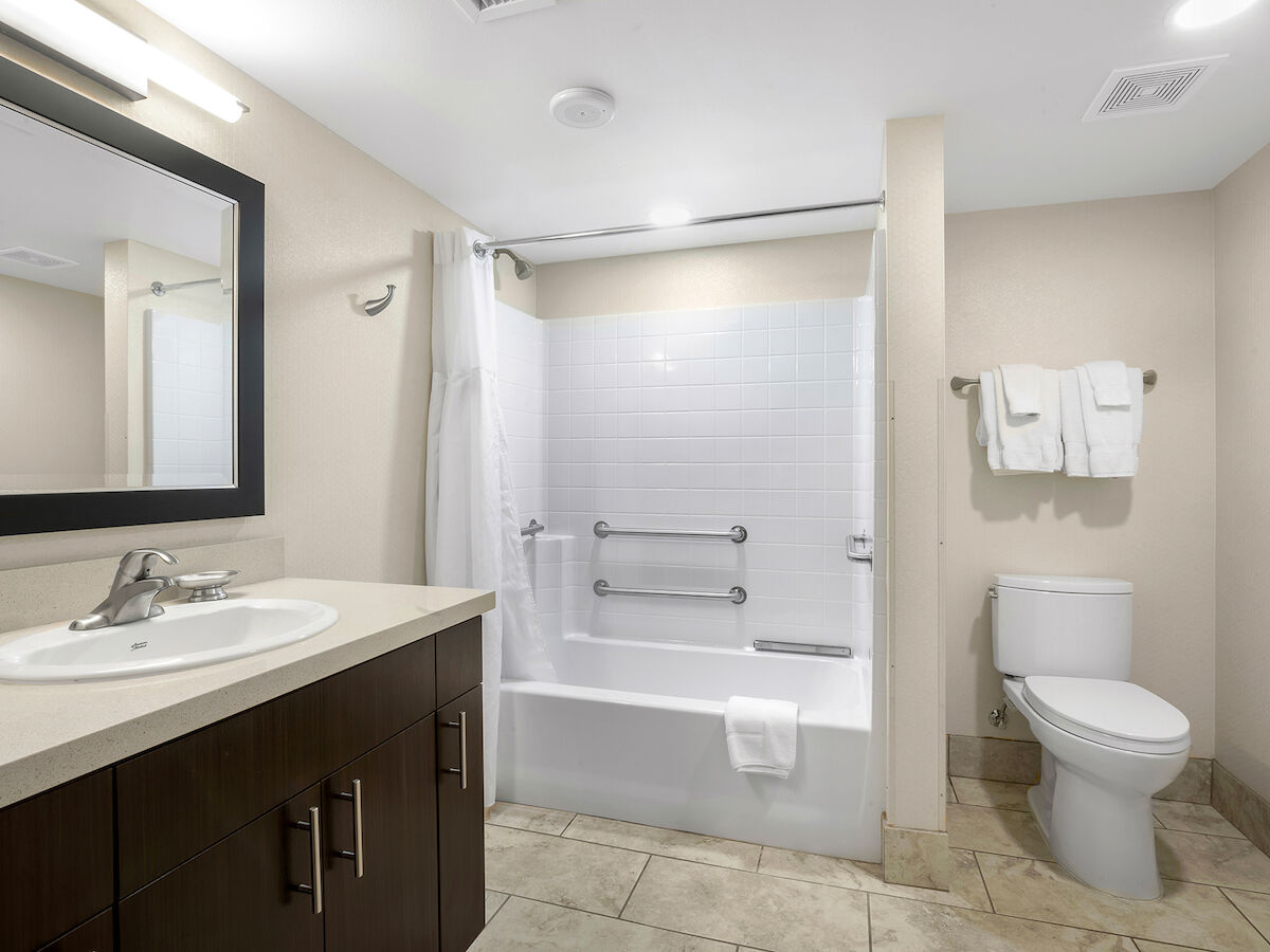 A clean and modern bathroom featuring a sink with a mirror, a bathtub with grab bars, white tiles, a toilet, and towels neatly hung on a rack.