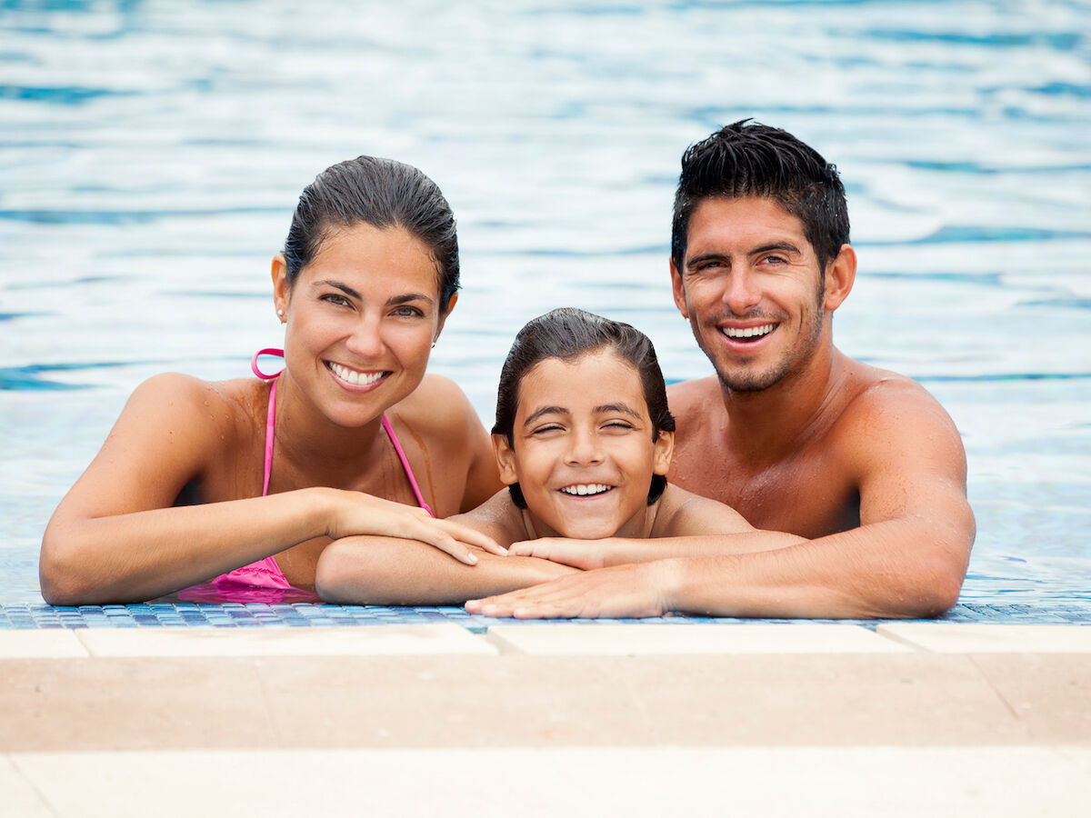 Three people are smiling while relaxing in a swimming pool, leaning on the edge with their arms resting on it.