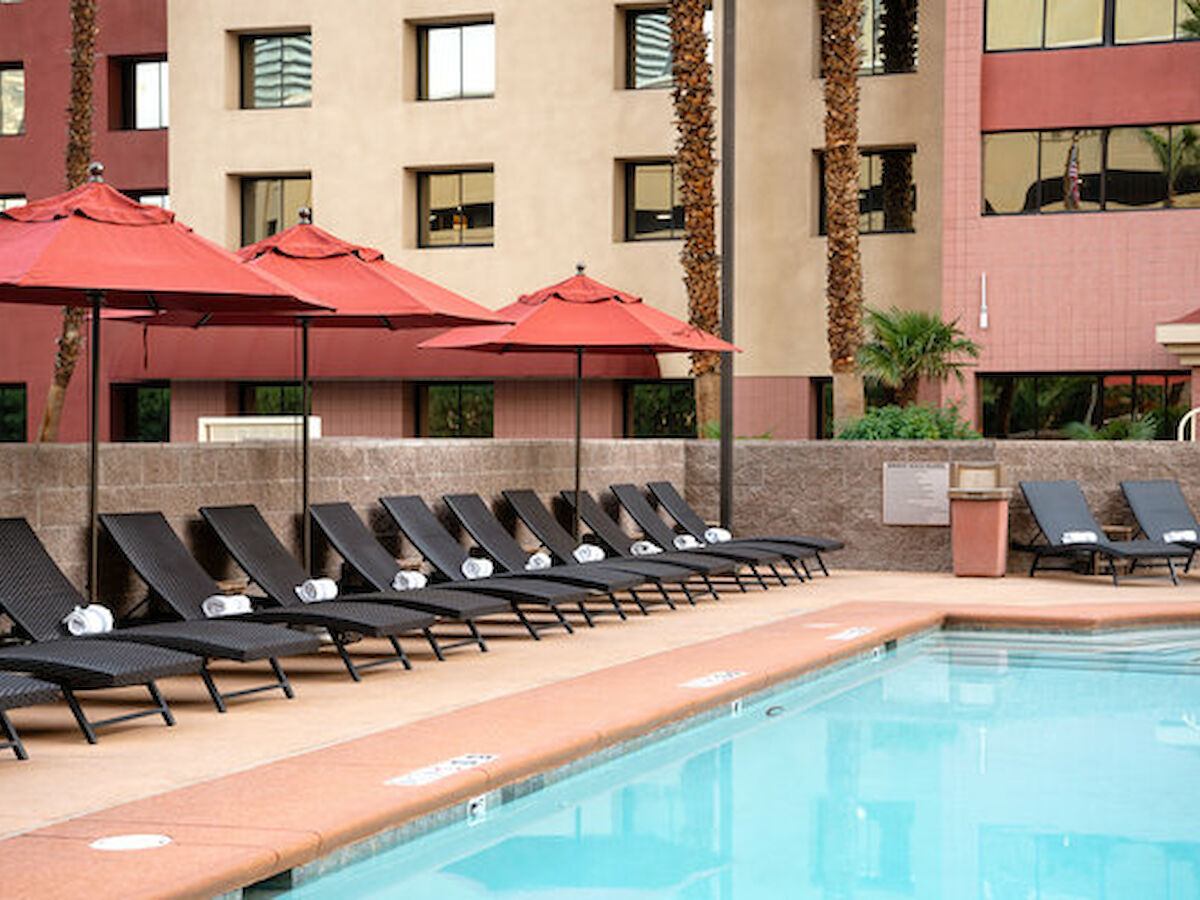 The image shows a poolside area with black lounge chairs, red umbrellas, neatly rolled towels, and a pool in front of an apartment building.