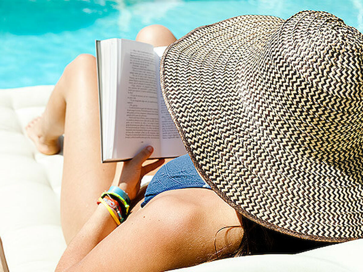 A person wearing a large sun hat is lying on a lounge chair by the pool, reading a book. They are enjoying a sunny day by the water.