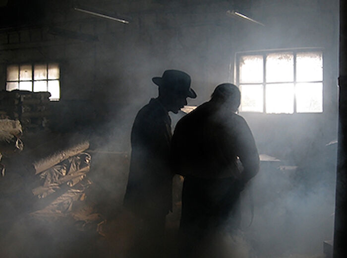 Two people in silhouette appear to be talking or working in a dimly lit, smoky room with stacks of materials and a window in the background.