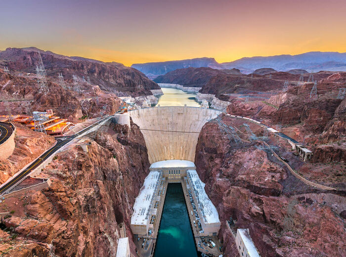 A massive dam stretches across a canyon, with a reservoir behind it and infrastructure at the base, set against a scenic mountainous backdrop at sunset.