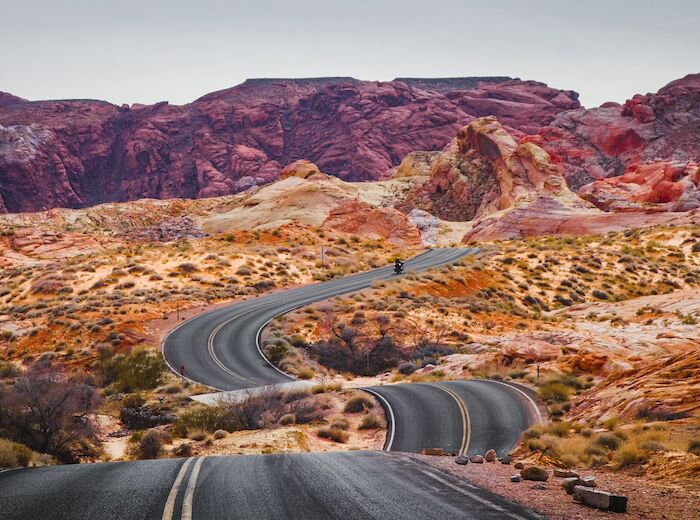 A winding road leads through a colorful desert landscape surrounded by rocky hills and sparse vegetation under a gray sky.