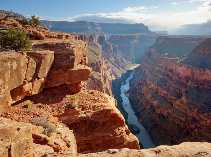 The image shows a breathtaking view of the Grand Canyon, with rugged cliffs and a river winding through the deep canyon under a partly cloudy sky.
