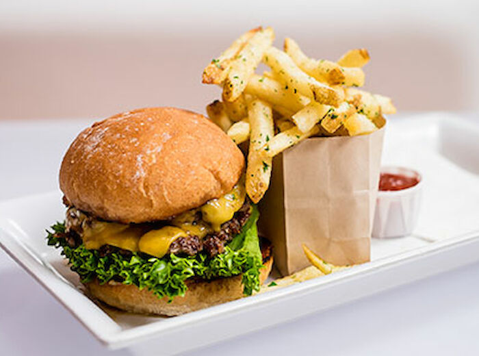The image shows a cheeseburger with lettuce and cheese, served with a side of French fries in a brown bag and a dish of ketchup on a rectangular plate.