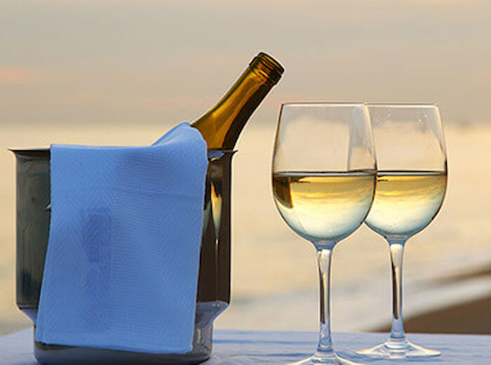 A wine bottle in an ice bucket with a blue cloth and two filled wine glasses on a table by the beach during sunset.