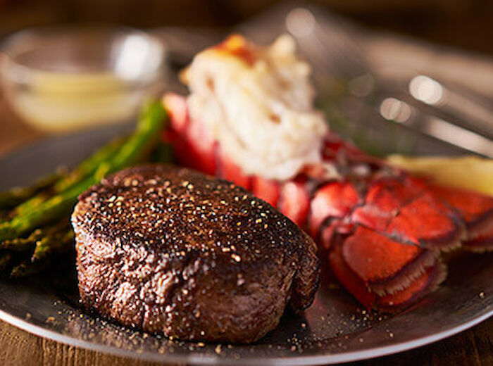 A plated meal featuring a grilled steak, lobster tail, and asparagus spears, with a small dish of melted butter in the background.