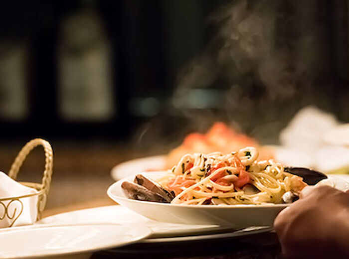 A hand is serving a steaming plate of pasta with seafood and vegetables, next to a bowl with a napkin and a heart-printed basket.