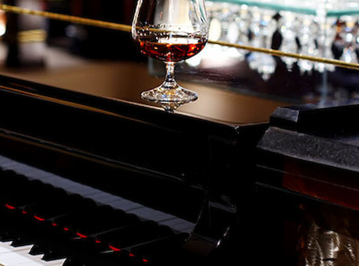 A wine glass sits on top of a black piano, with its keys and a reflective surface visible, likely in a bar or elegant setting.