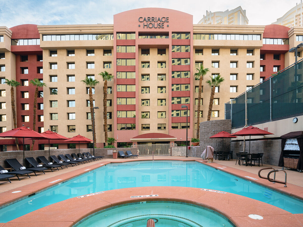 The image shows a hotel building with the sign "Carriage House," a swimming pool in the foreground, lounge chairs, and umbrellas. Guests can relax here.