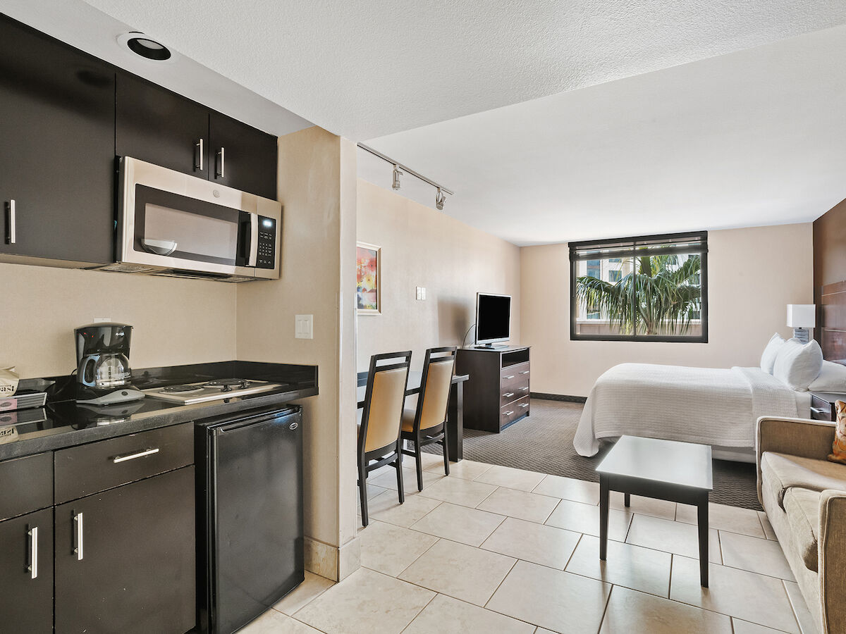 A modern hotel room features a kitchenette, seating area, queen-size bed, desk, TV, dresser, and a large window with a view.