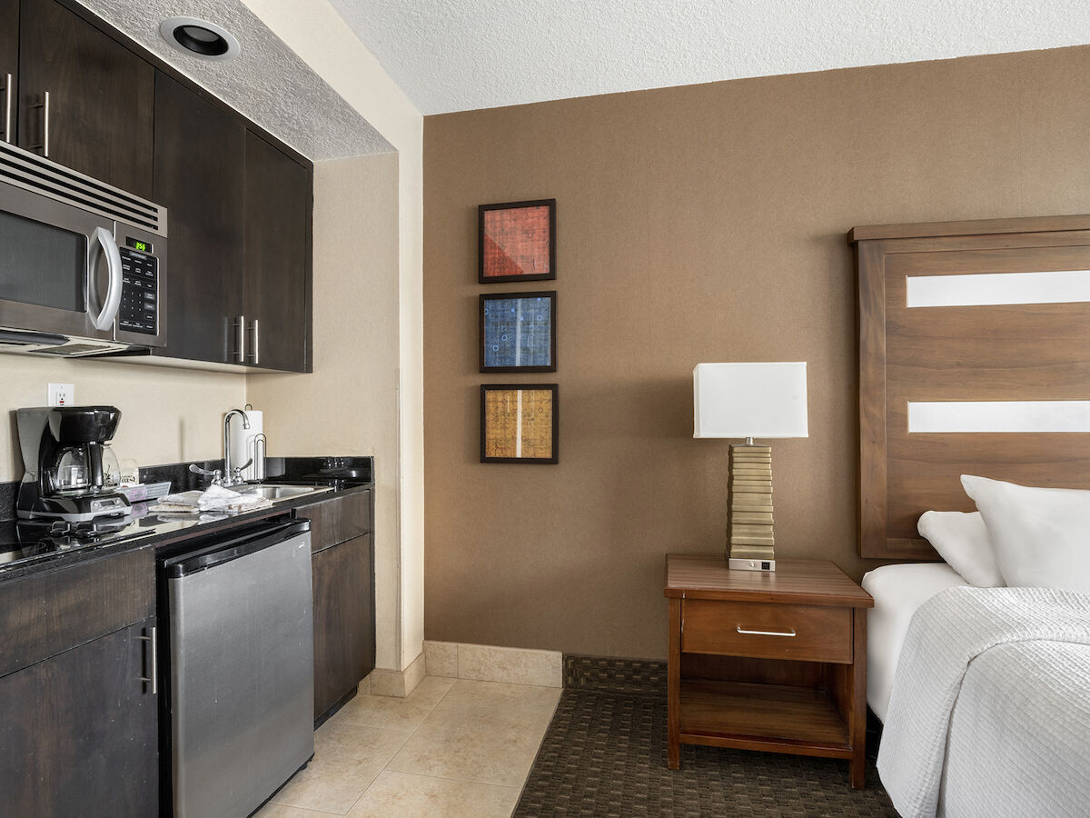 The image shows a hotel room with a kitchenette, a bed, a nightstand with a lamp, and three framed artworks on a brown accent wall.