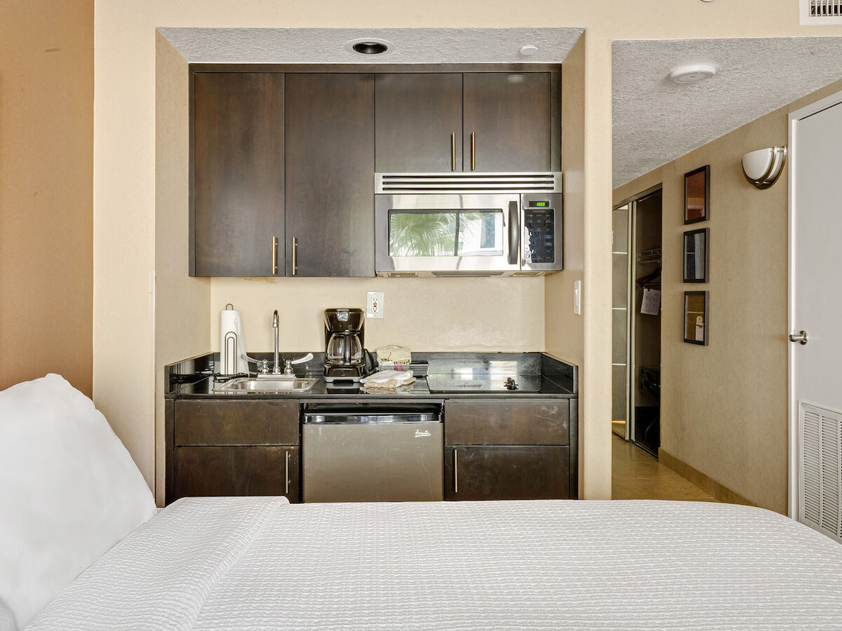 A compact studio or hotel room features a bed, kitchenette with microwave, mini-fridge, sink, cabinets, and a hallway leading to a closed door.