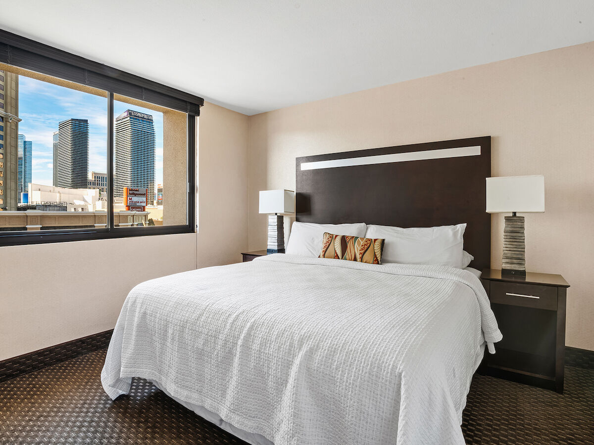 A modern bedroom with a large bed, two bedside tables with lamps, and a window offering a view of city buildings.