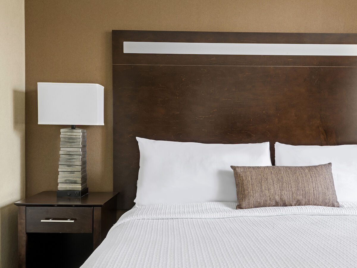 The image shows a neatly made bed with white pillows and a brown accent pillow, next to a nightstand with a modern lamp on it.