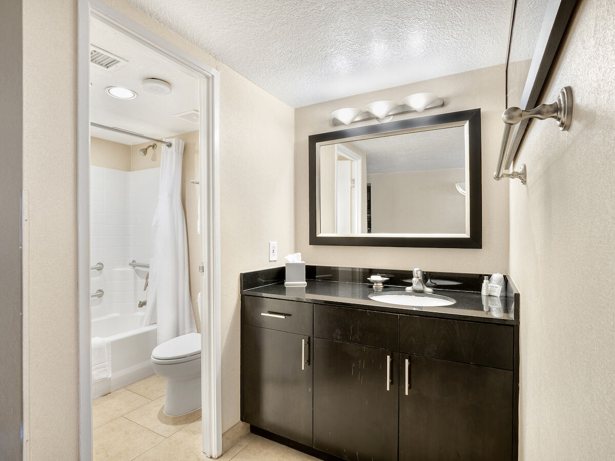 A clean bathroom featuring a large mirror, sink with a dark cabinet, well-lit vanity, and a toilet with a shower in the adjacent room ending the sentence.