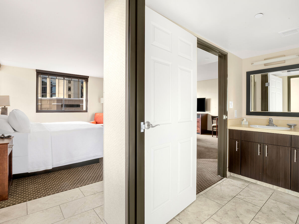 The image shows a hotel room with a bed, a bedside table, a bathroom vanity with a sink, and an open door leading to a living area.