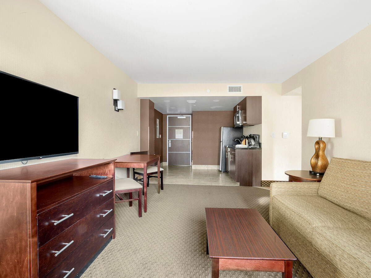 A hotel room with a TV, a sofa, a coffee table, a desk, and chairs. The kitchenette area includes a fridge, microwave, and cabinets.