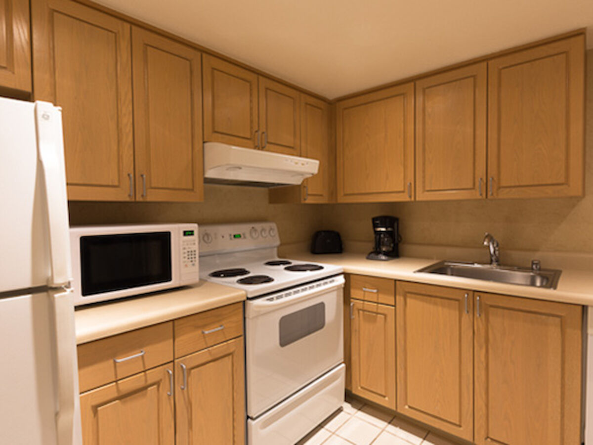 A small kitchen with wooden cabinets, a white refrigerator, microwave, stove, toaster, coffee machine, and a stainless steel sink.
