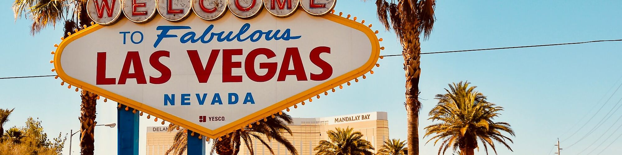 The image shows the iconic "Welcome to Fabulous Las Vegas Nevada" sign surrounded by palm trees under a clear blue sky, with a view of the city.