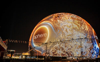 A large illuminated spherical structure displays the word "sphere" against a night sky, with surrounding scaffolding and buildings visible in the foreground.