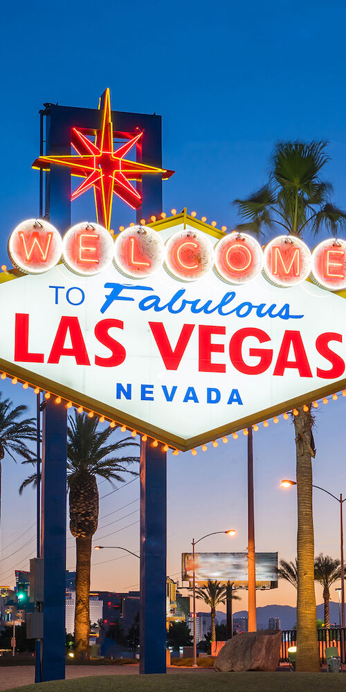 The image shows the iconic "Welcome to Fabulous Las Vegas, Nevada" sign at dusk, surrounded by palm trees, with the cityscape in the background.