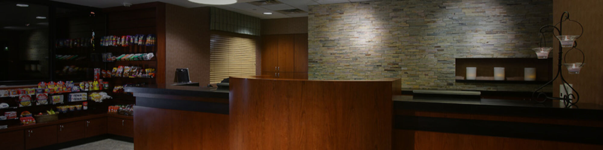 The image shows a reception desk area with wooden finishes and stone walls, featuring modern lighting and a snack display shelf to the left.