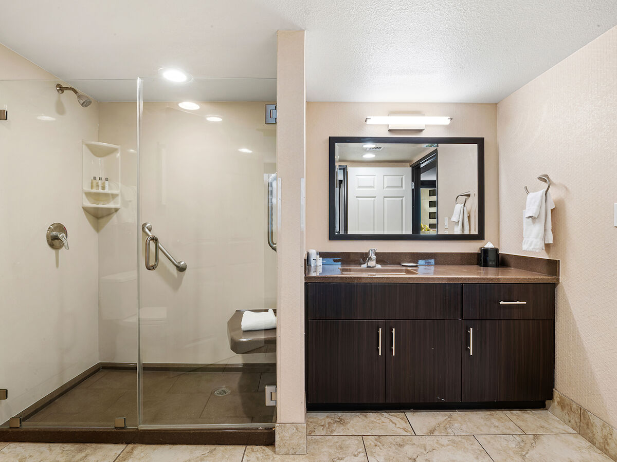 The image depicts a modern bathroom with a glass-enclosed shower, a dark wood vanity with a rectangular mirror above it, and beige tiled flooring.