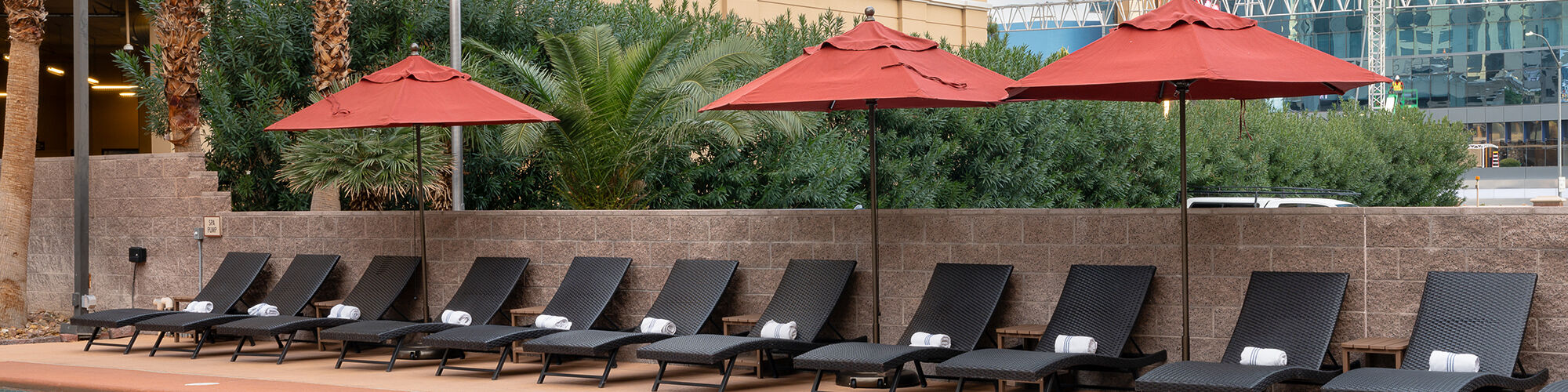 A poolside area with lounge chairs and red umbrellas, adjacent to a parking structure and surrounded by greenery.
