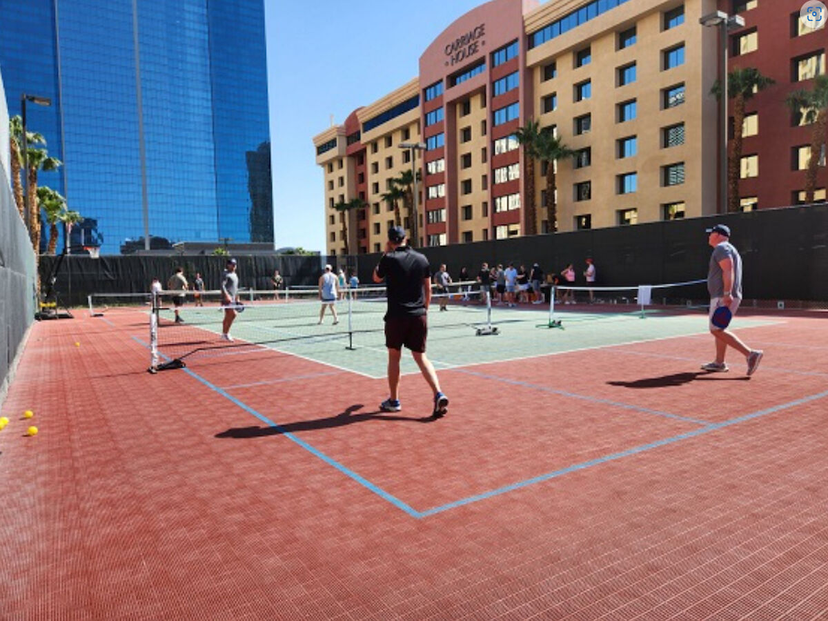 People are playing pickleball on a rooftop court surrounded by buildings, with a group watching nearby. The weather appears to be sunny.